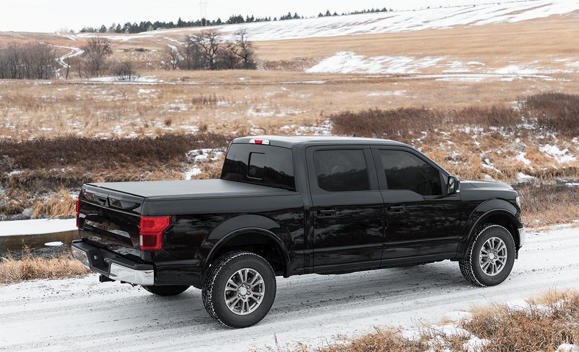 A black truck equipped with a roll-up tonneau cover navigating a snowy road in a winter setting.