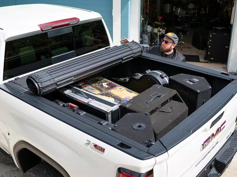A man sits in the back of a truck with a toolbox, showcasing the BAK Revolver X4S Tonneau Cover above him.
